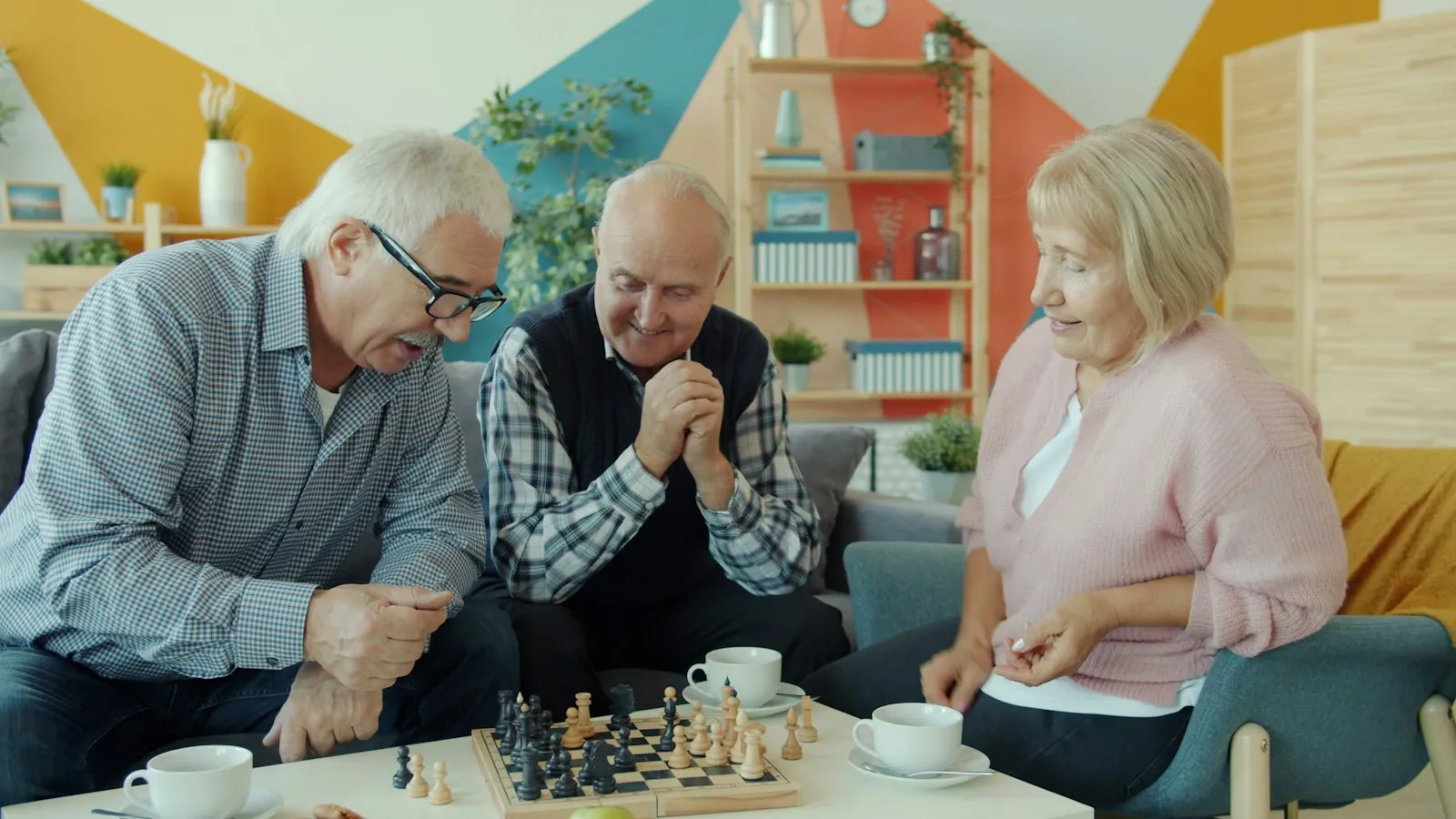 Two older men laughing over a game of chess while a friend looks on
